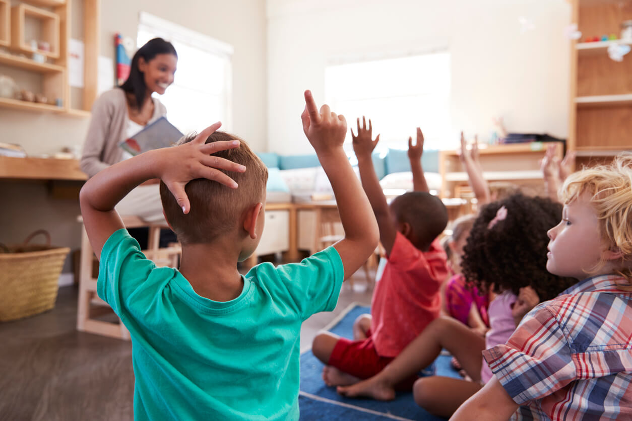 A group of children sit on the floor, raising hands, with a person reading to them in a bright classroom setting.