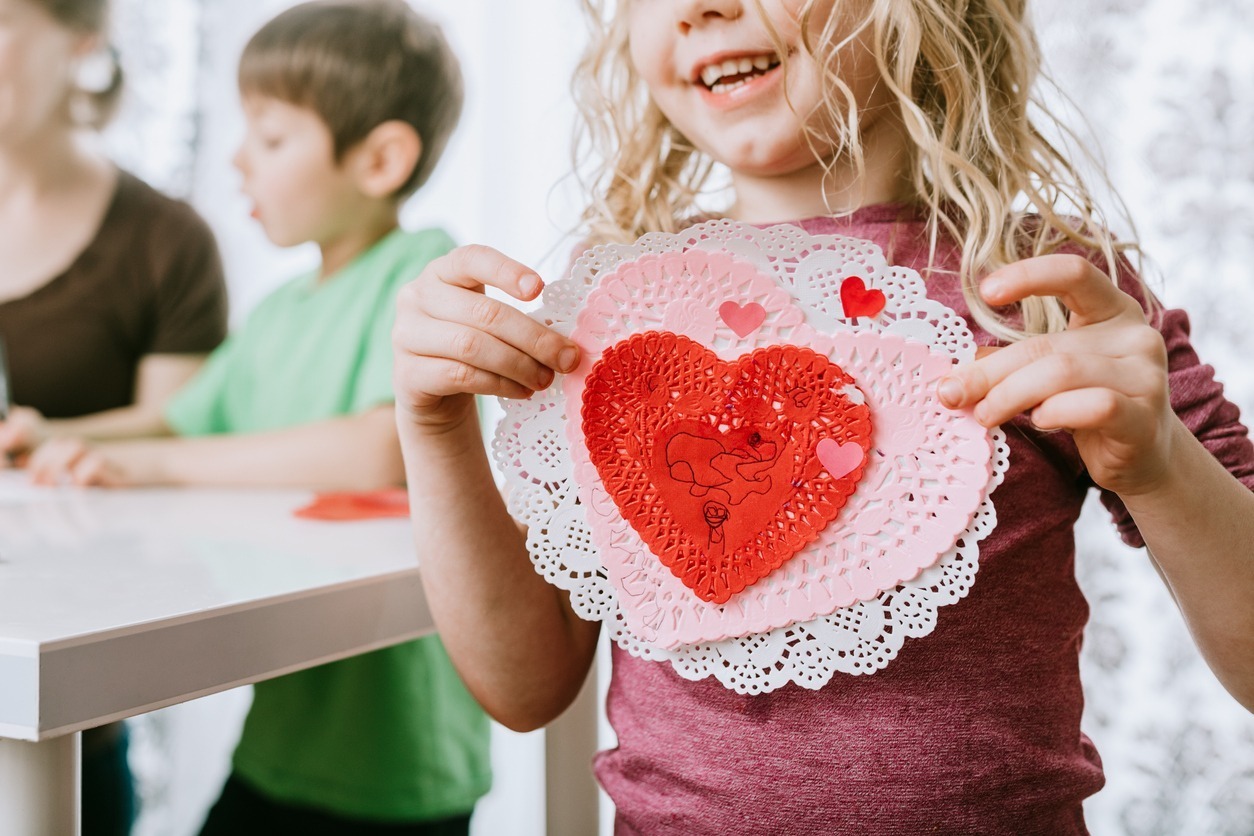 A child holds a handmade heart craft while smiling. Another child and a person are in the background, sitting at a table.