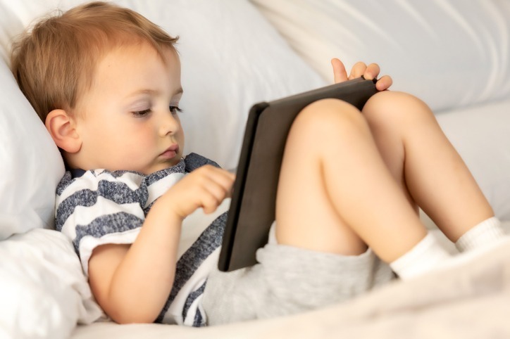 A young child in striped shirt and shorts reclines on white bedding, attentively using a tablet in a relaxed, comfortable setting.