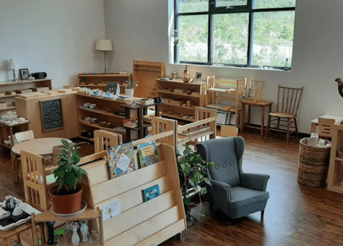 A cozy, well-lit classroom with wooden furniture, shelves of books and plants, a grey armchair, and various learning materials neatly arranged.