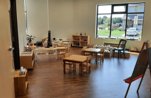 Bright classroom with wooden tables, chairs, and shelves. Large window showing greenery outside. Art supplies and learning materials neatly arranged inside.