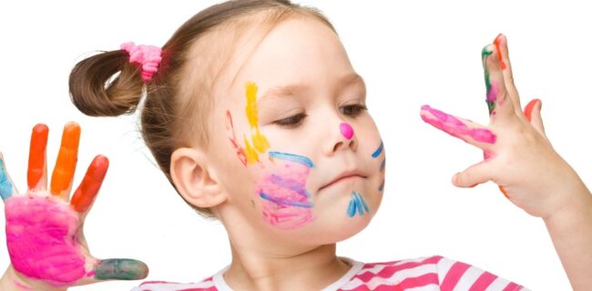 A child with colorful paint on their face and hands, wearing a striped shirt, looks at painted fingers against a plain background.