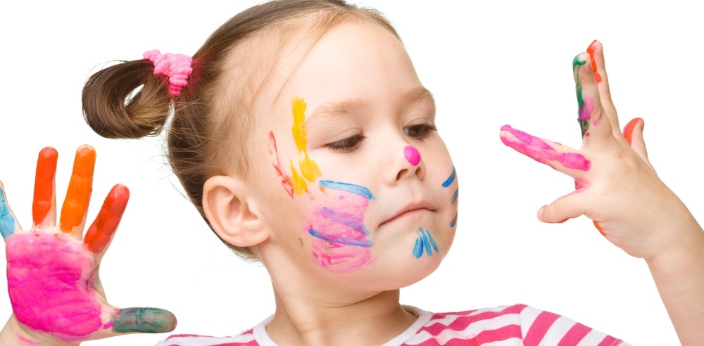 A child with colorful paint on their face and hands, wearing a striped shirt, looks at painted fingers against a plain background.