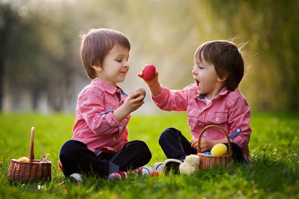 Two young children sitting on grass, wearing red shirts, happily playing with colorful eggs and baskets, in a sunny, green outdoor setting.