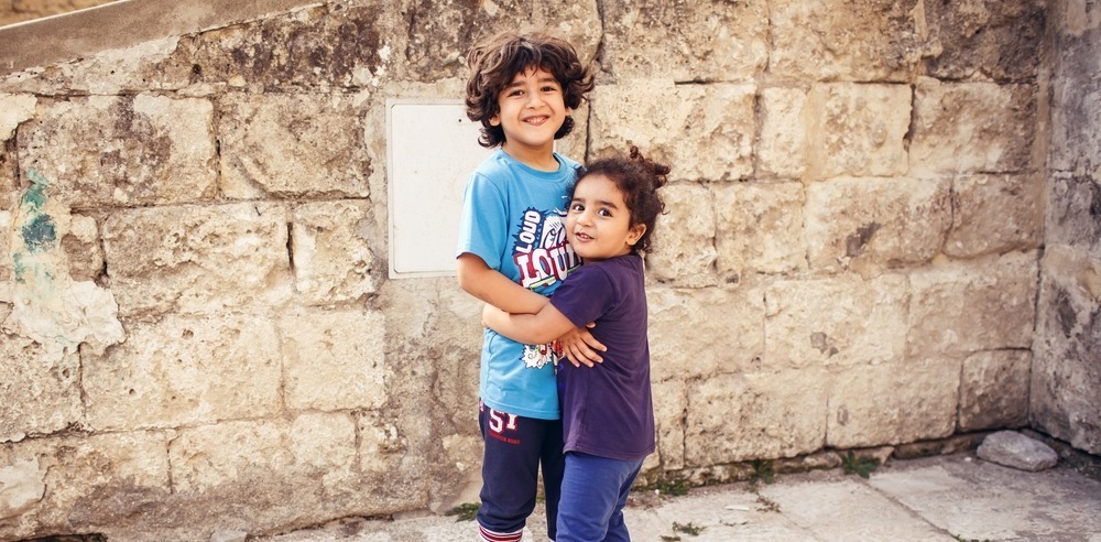 Two children smiling and hugging in front of an aged stone wall. Their playful and warm expressions create a heartwarming and joyful scene.
