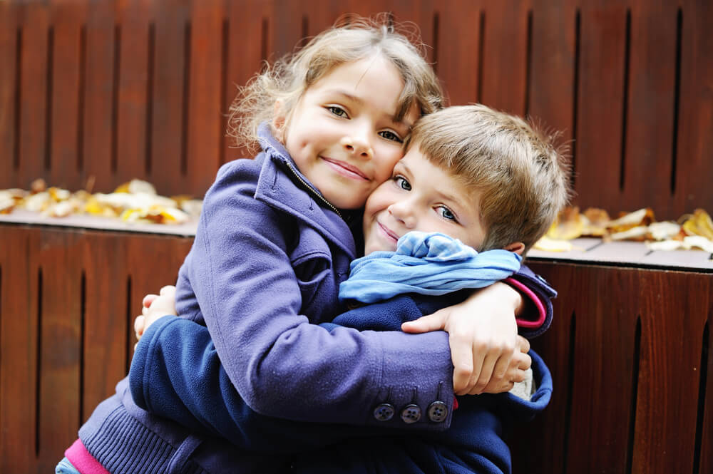 Two children wearing coats hugging joyfully in front of a wooden bench, surrounded by fall leaves. Warm, affectionate moment captured outdoors.