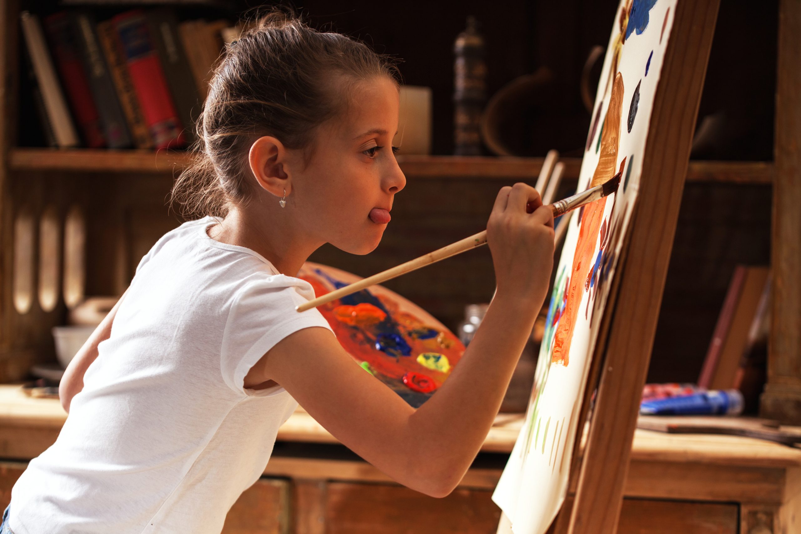 A young child in a white shirt is focused on painting with vibrant colors, surrounded by books and art supplies.