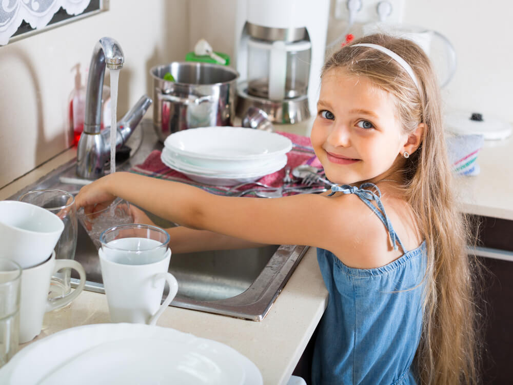 A child with long hair washes dishes at a kitchen sink, smiling. Various kitchenware and a faucet are visible in the background.