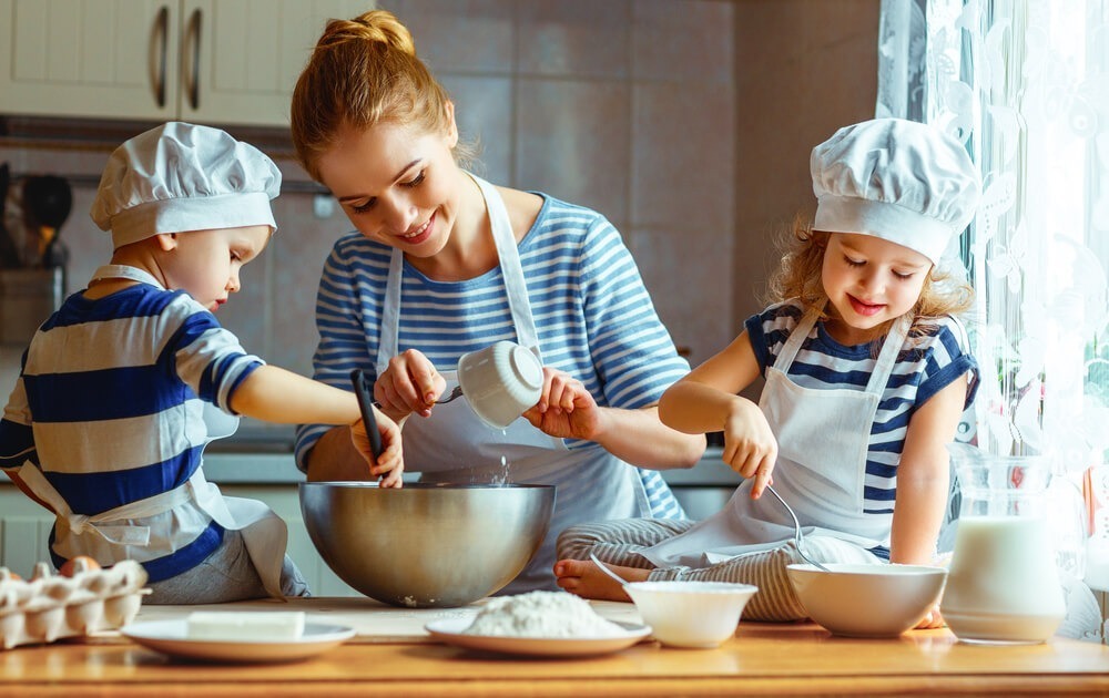 A person and two children in matching outfits bake together, smiling and mixing ingredients in a kitchen setting.