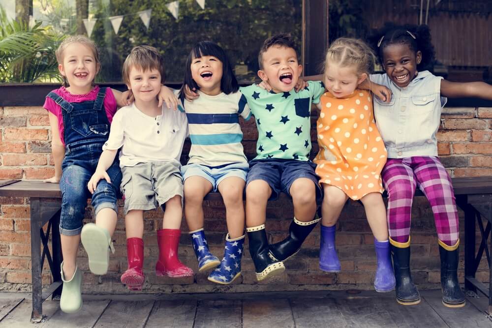 Six children sit joyfully on a bench, wearing colorful outfits and boots, with a brick wall and festive bunting in the background.