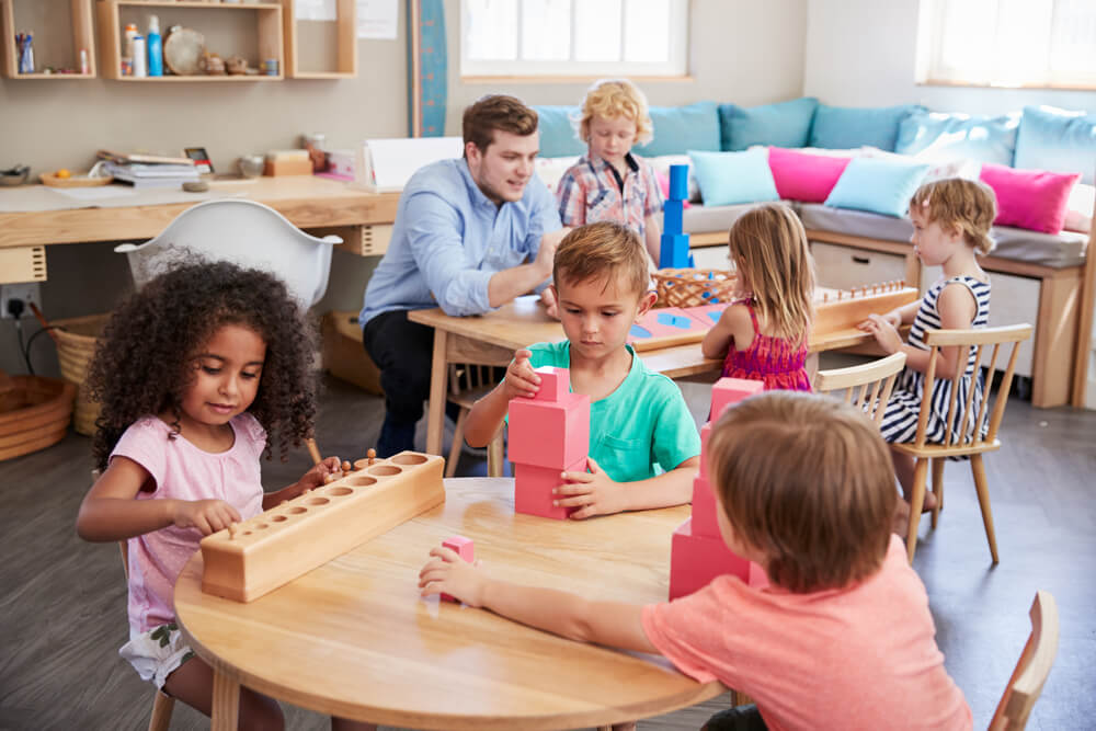 Children play with educational toys in a classroom, where a person assists them. Bright cushions are visible on a seating area.
