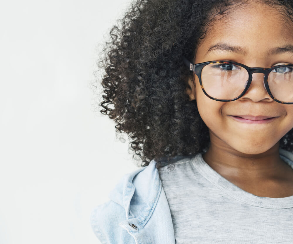 A young child with curly hair and glasses smiles warmly at the camera, wearing a gray shirt and light blue jacket.