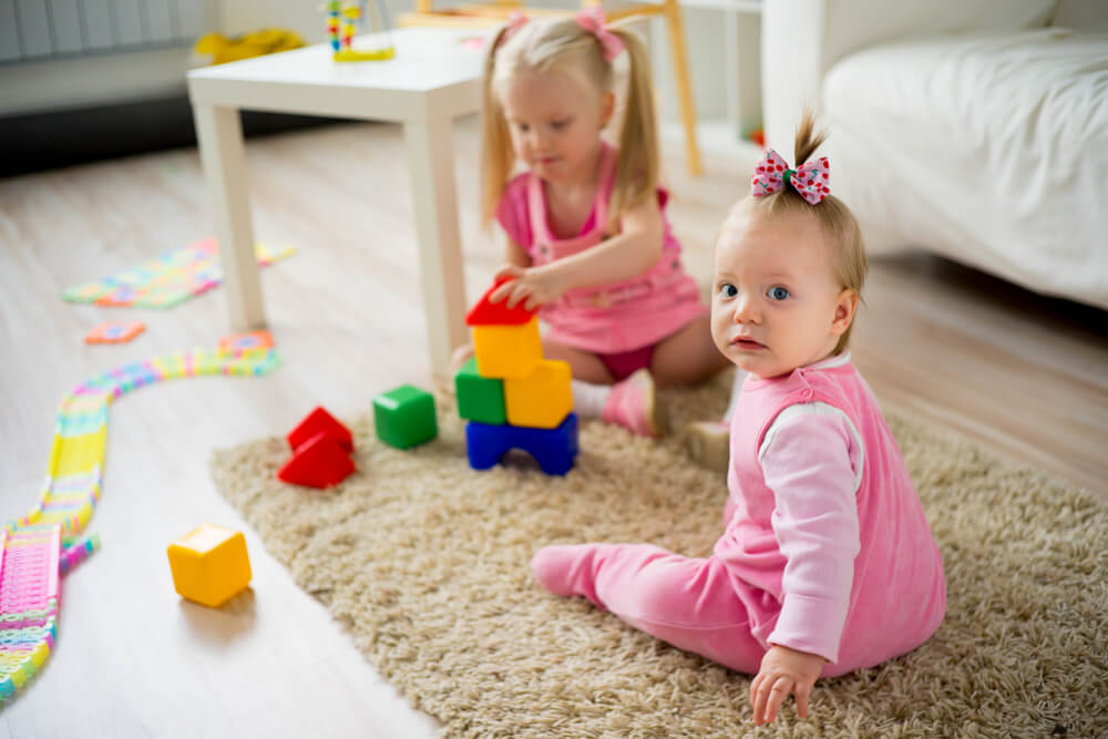Two young children play with colorful blocks on a carpeted floor in a bright, cozy room with toys and a couch visible.