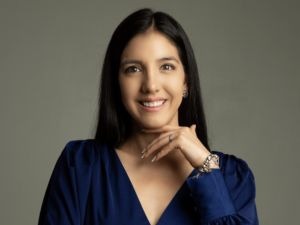 A person with long dark hair and a blue top smiles against a plain background, resting their chin on their hand.