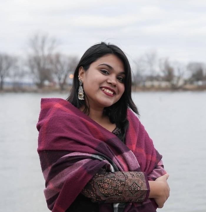 A person wearing a patterned outfit and shawl smiles near a body of water, with bare trees in the background under a cloudy sky.