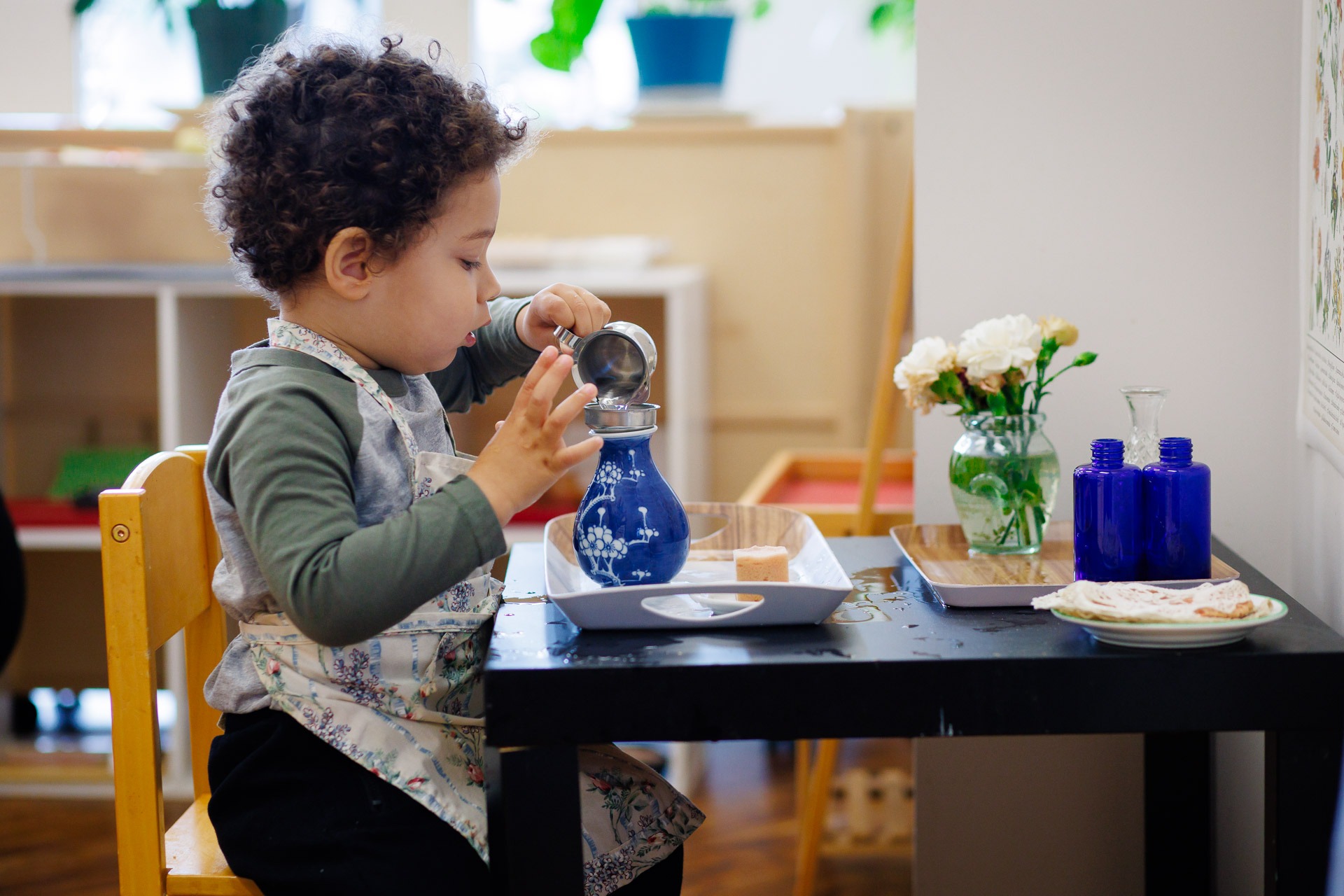 A child carefully pours water into a decorative blue vase, sitting at a small table with flowers and blue bottles nearby.