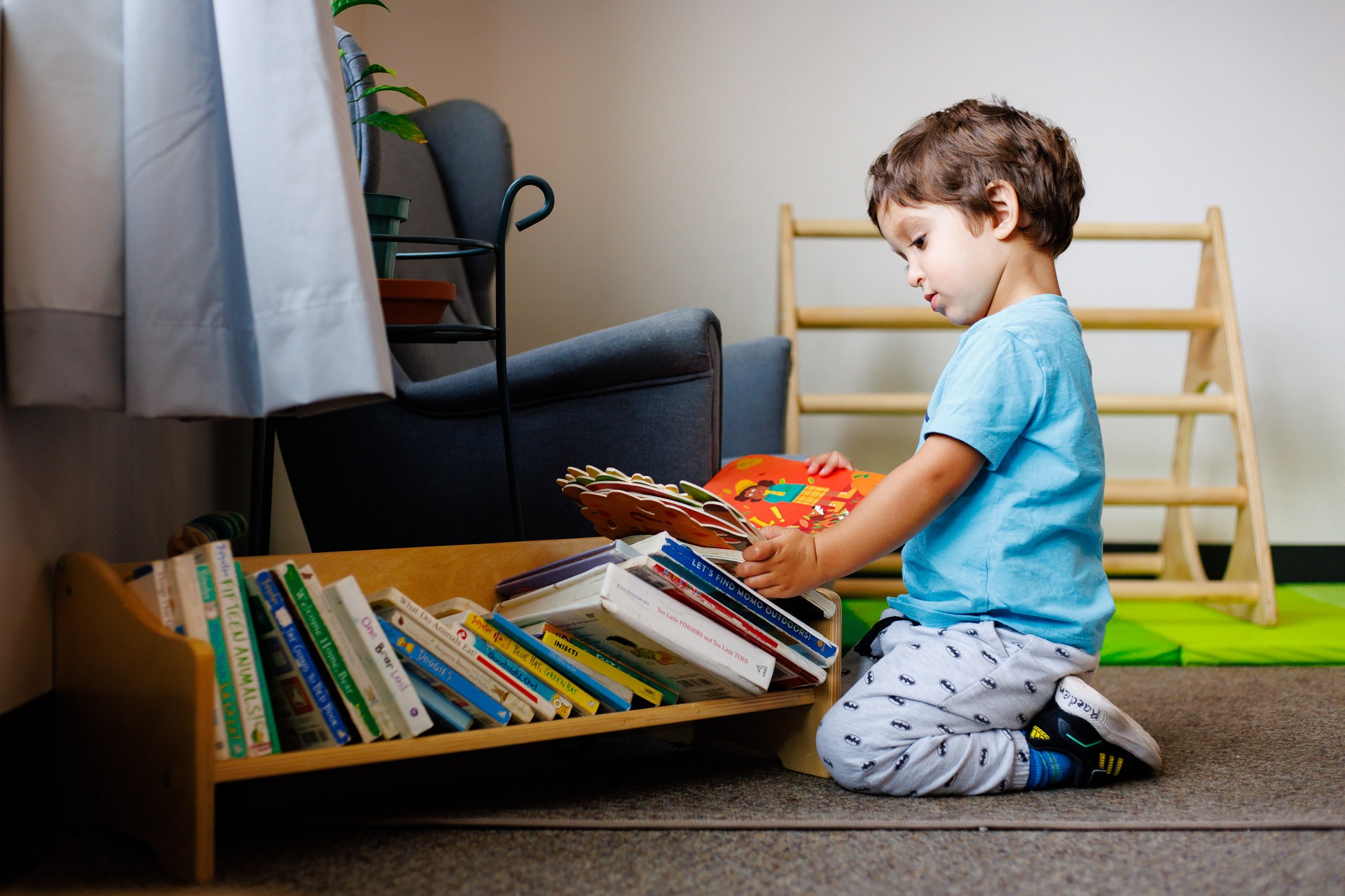 A child in a blue shirt selects a colorful book from a low bookshelf in a cozy, well-lit room.