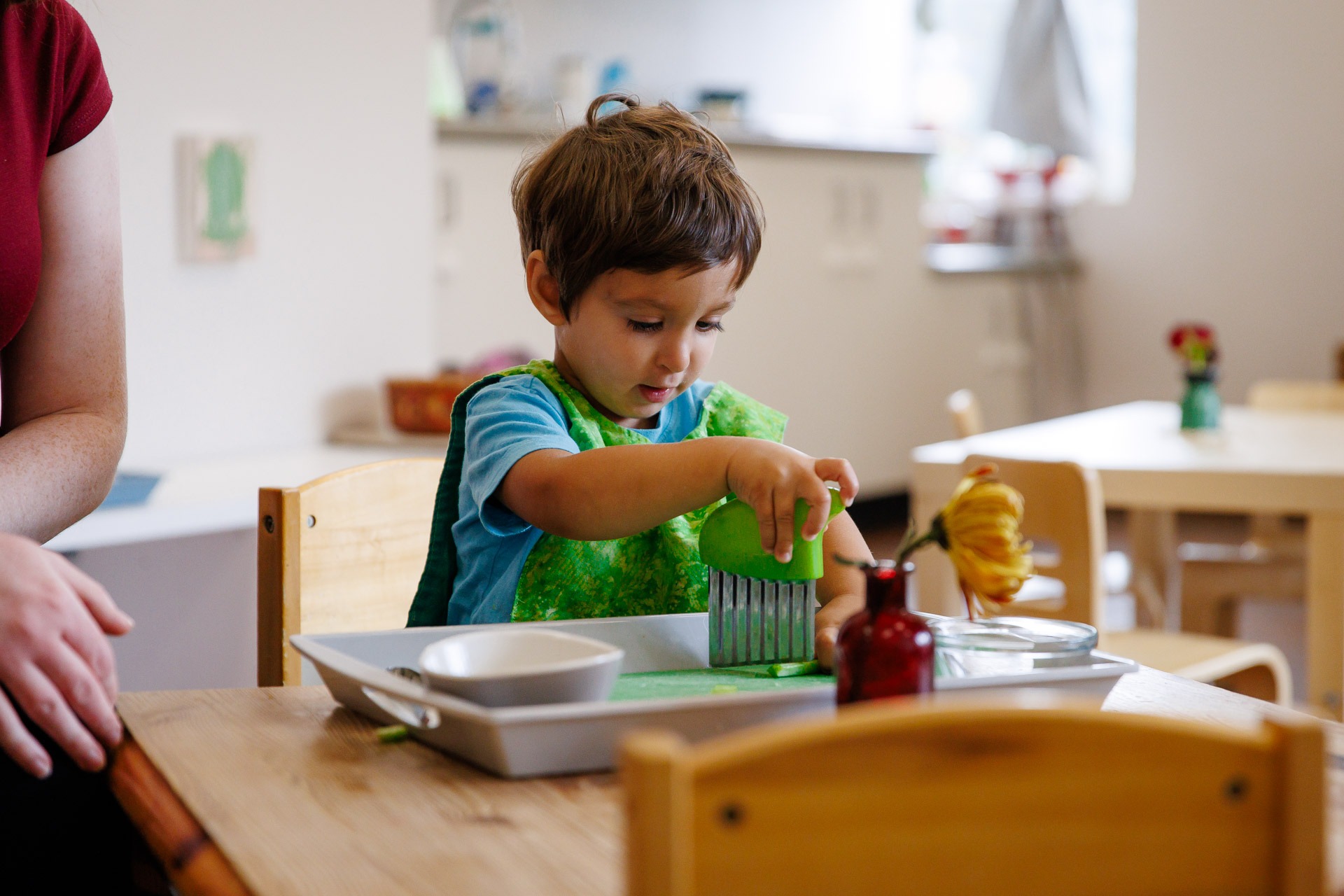 A child plays in a classroom setting with a green tool, while a person sits nearby, surrounded by wooden furniture.