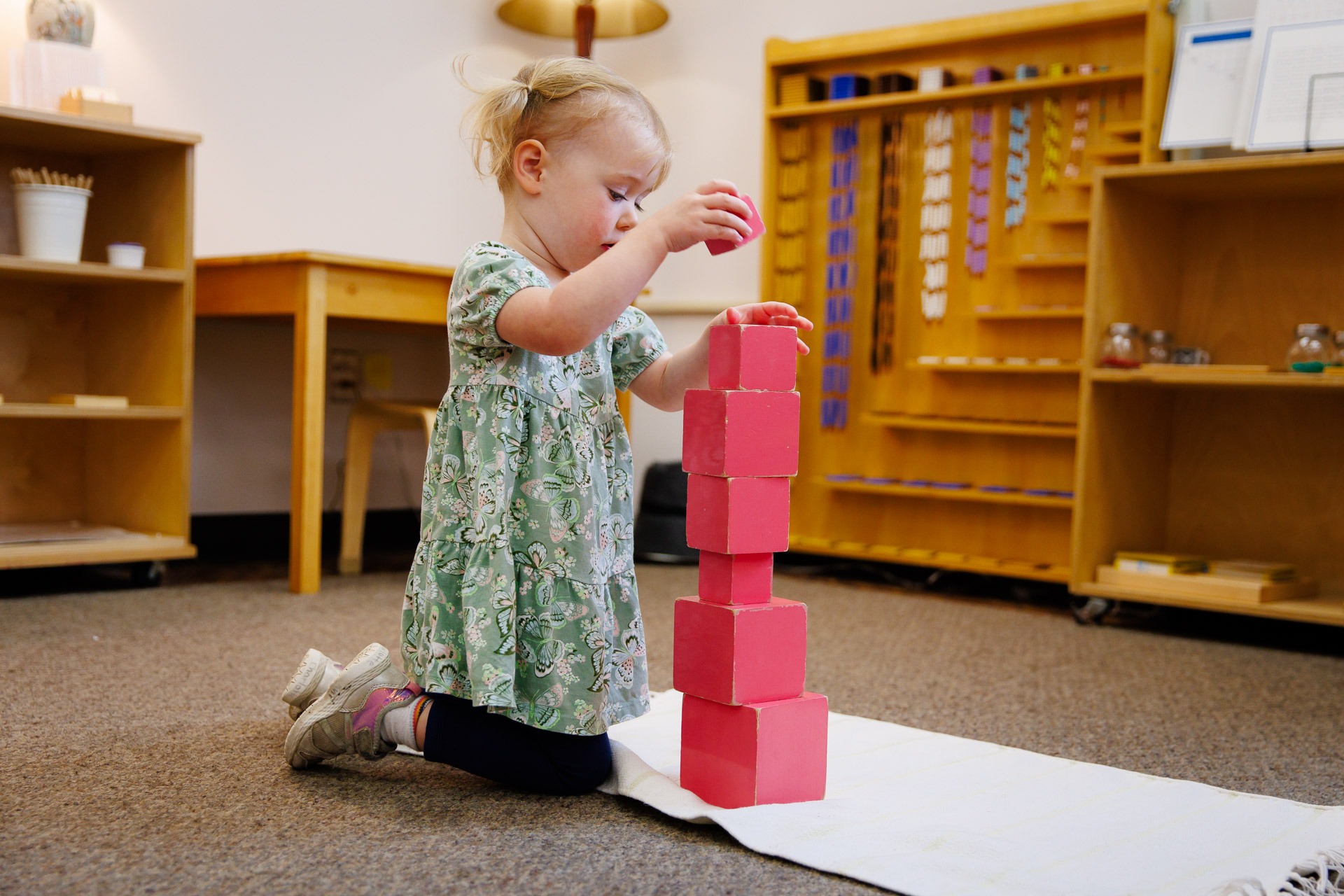 A child stacks red blocks on a white mat in a classroom with wooden shelves and educational materials in the background.