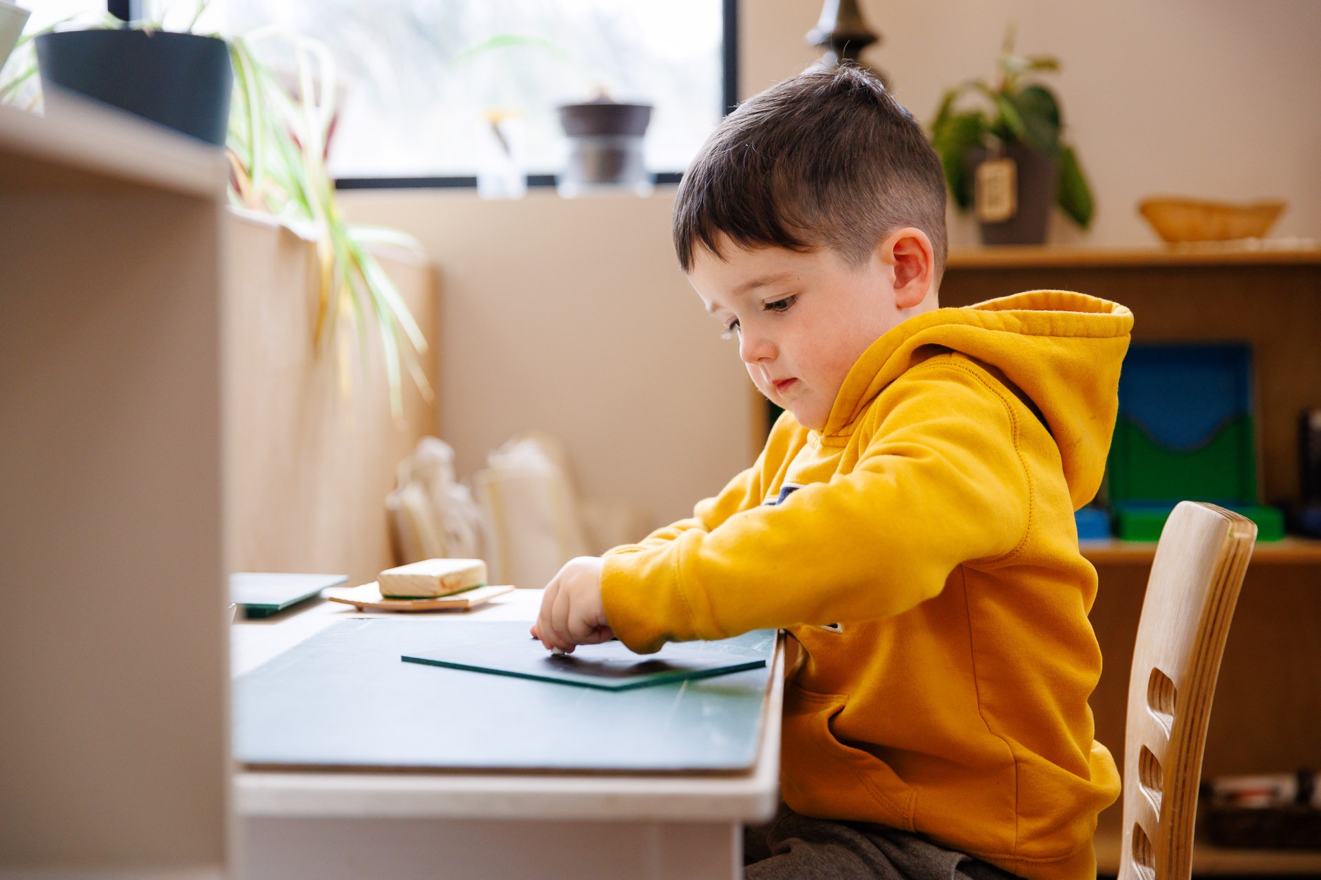 A child in a yellow hoodie is focused on a craft project at a table in a bright, plant-decorated room.