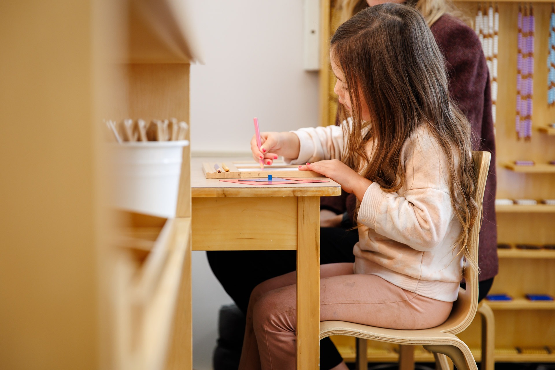A child is drawing at a wooden table, while a person sits nearby. Shelves with educational materials surround them in a classroom.