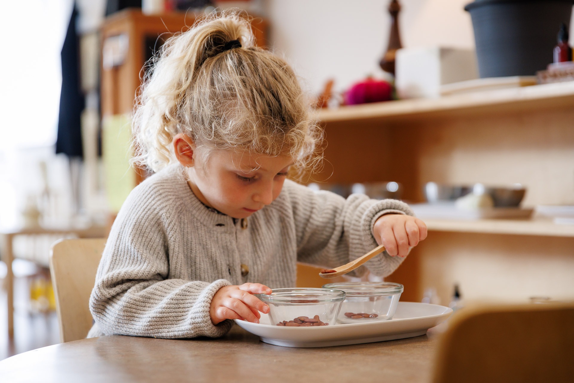 A child in a cozy sweater carefully transfers small items between glass bowls with a wooden spoon, focused on the task.