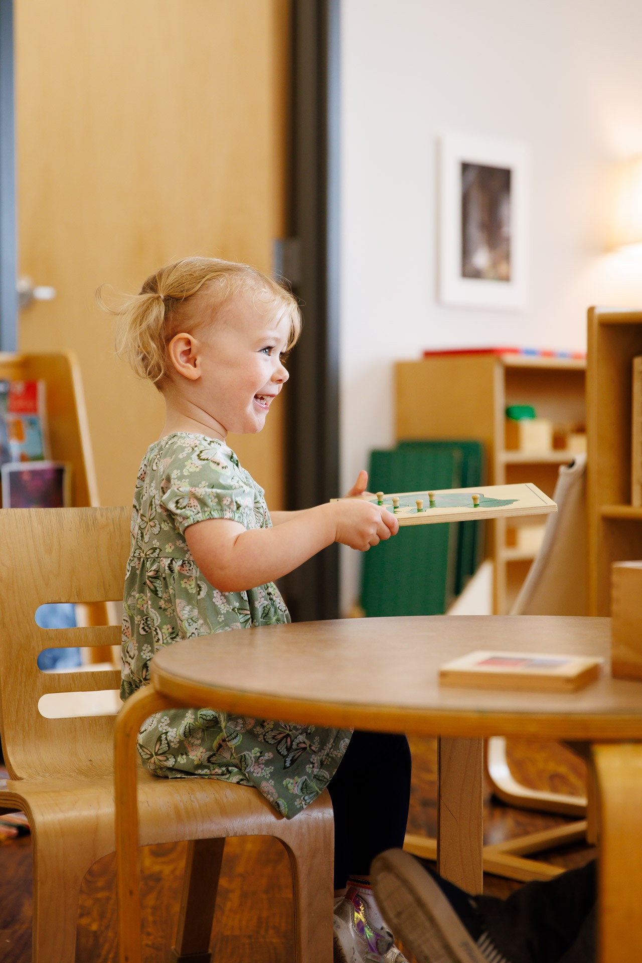 A child sits at a wooden table, holding a puzzle piece, surrounded by shelves and a painting in a warmly lit room.