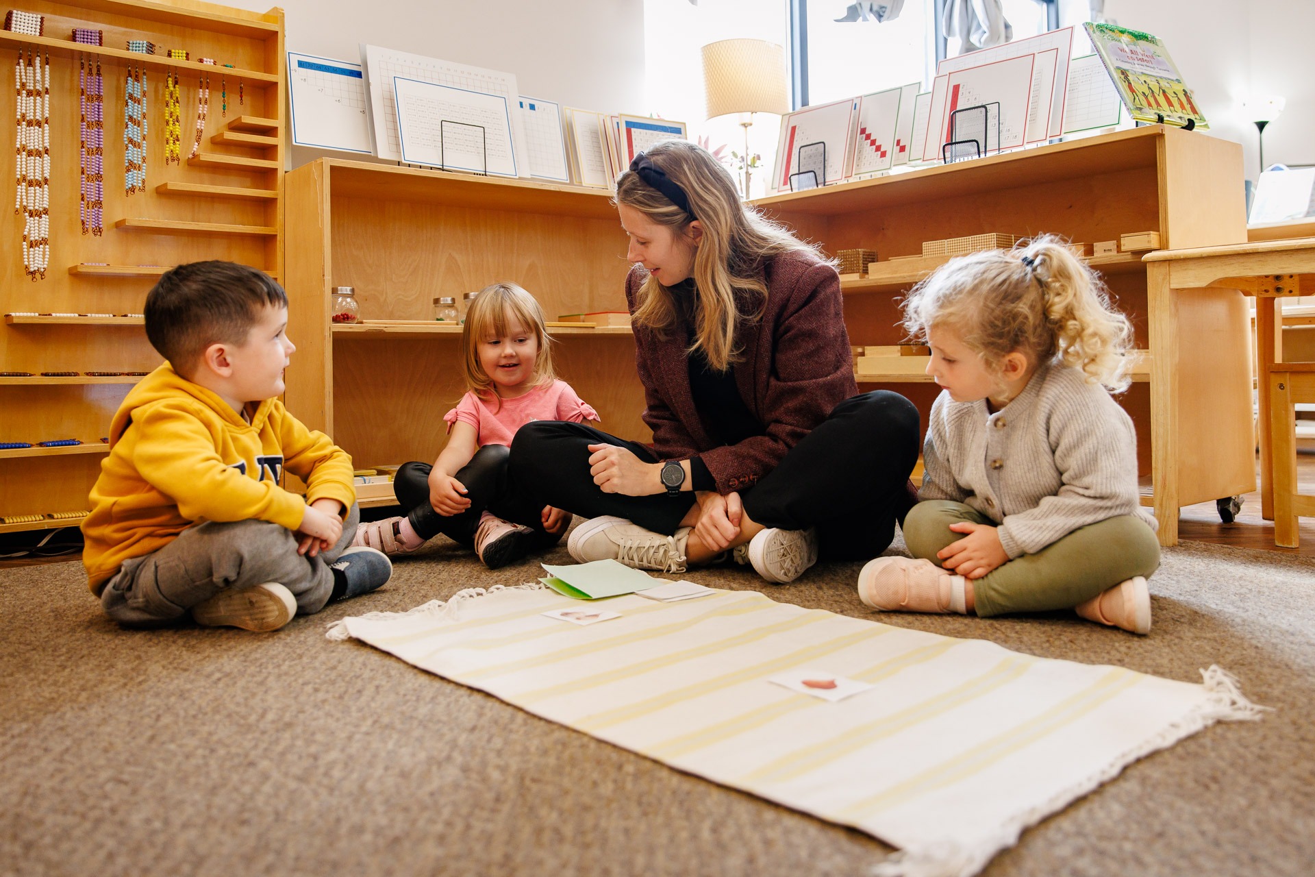 A person and three children sit on the floor in a classroom, engaged in an educational activity with materials.