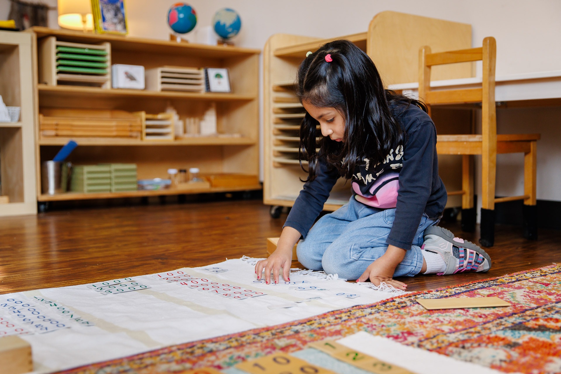 A child engages in a number activity on the floor, surrounded by wooden shelves and educational materials in a classroom setting.