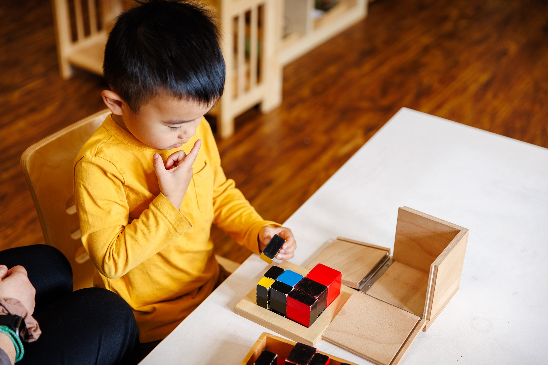 A child in a yellow shirt plays with colorful blocks at a table in a warmly lit classroom, next to a person.