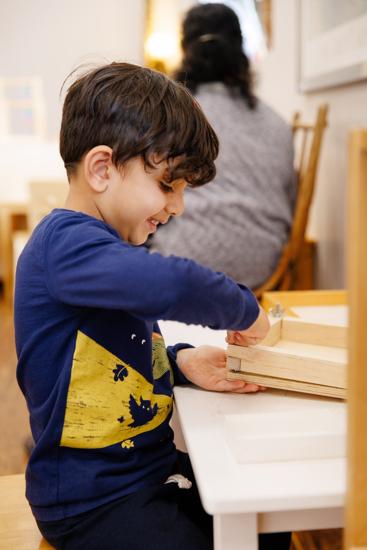 A child enthusiastically works on a wooden project while a person is seated nearby, in a cozy, well-lit room.