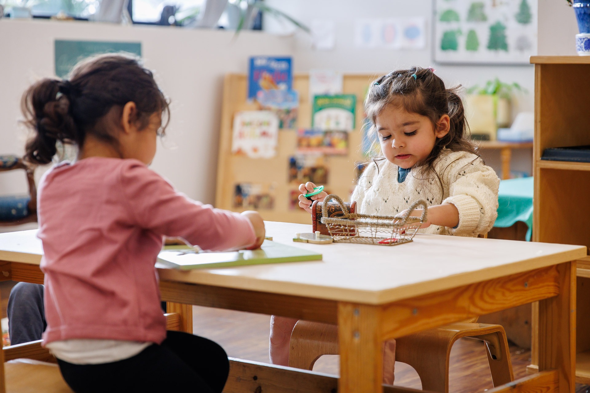 Two children engage in activities at a small table in a classroom setting, surrounded by educational materials and books.
