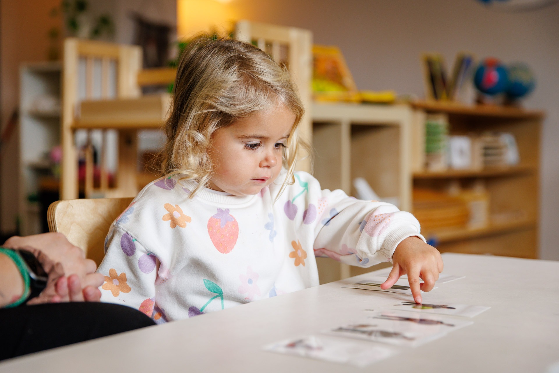 A child in a colorful sweater is pointing at cards on a table in a cozy, educational setting. Shelves with books are behind.