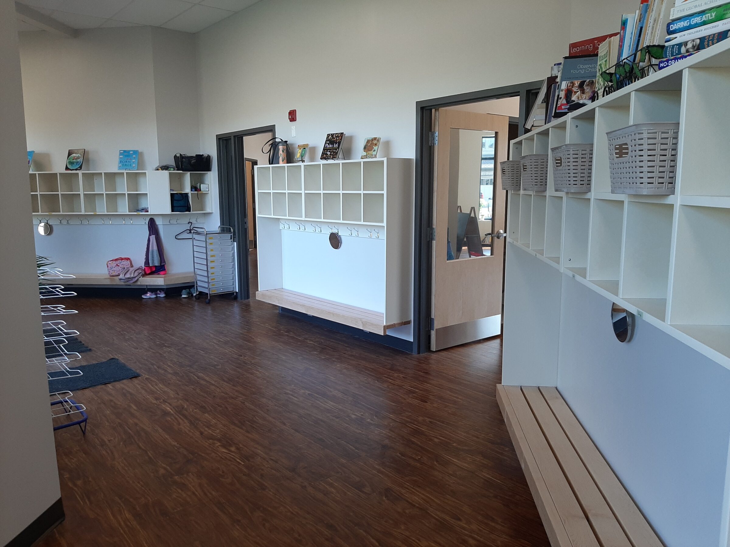 The image shows an empty school hallway with cubbies, books, a bench, and coat racks. A door opens to another classroom area.