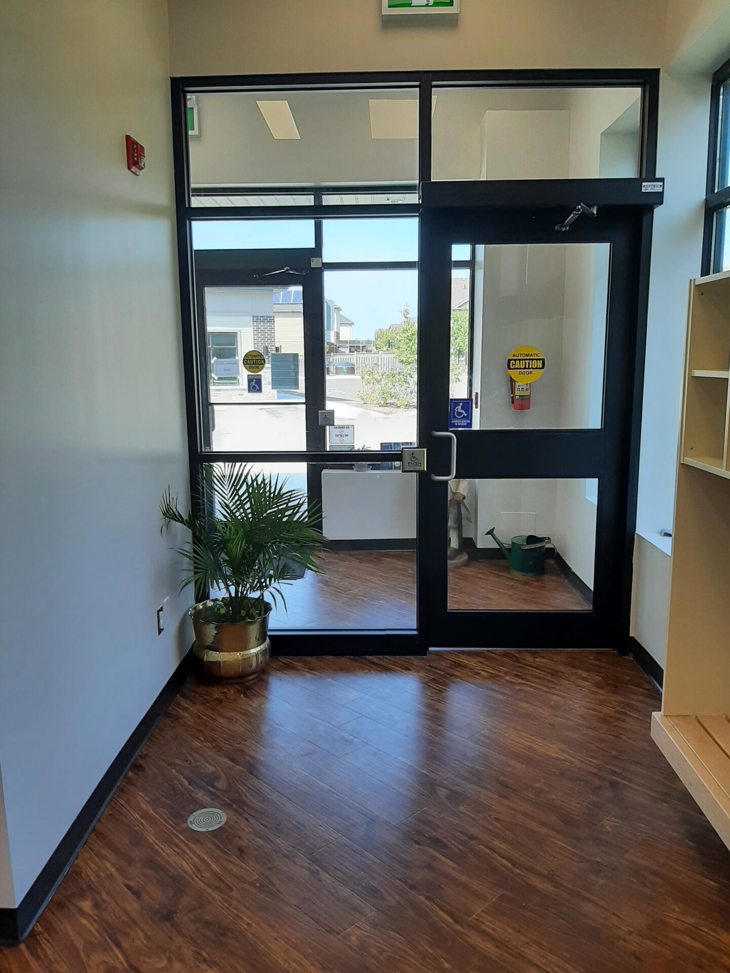 A foyer with wooden flooring features glass doors, a potted plant, and exit signs. A fire alarm and wheelchair sticker are visible.
