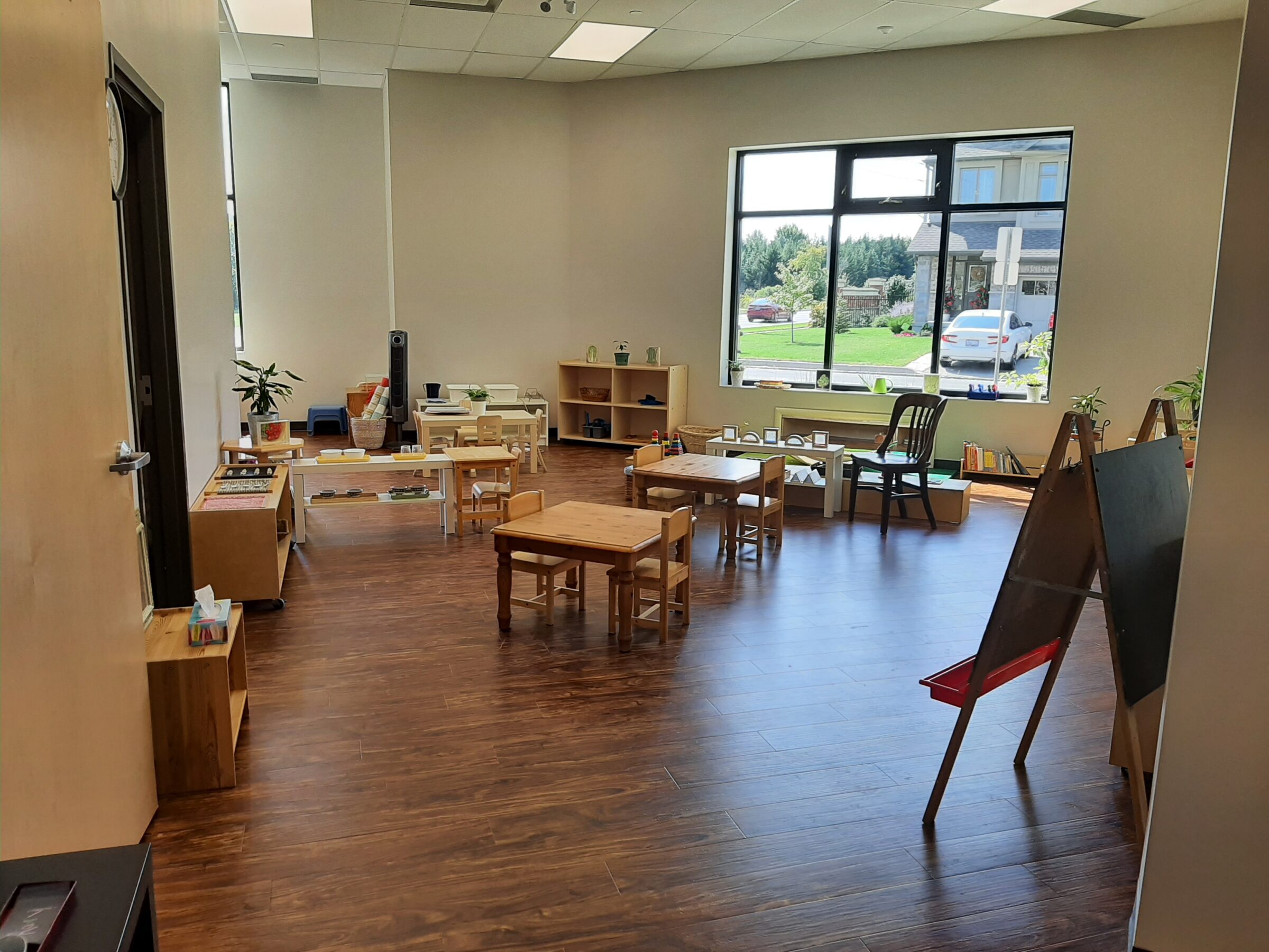 Bright classroom with wooden floors, small tables and chairs, educational toys, and plants. Large windows offer views of outside greenery and cars.