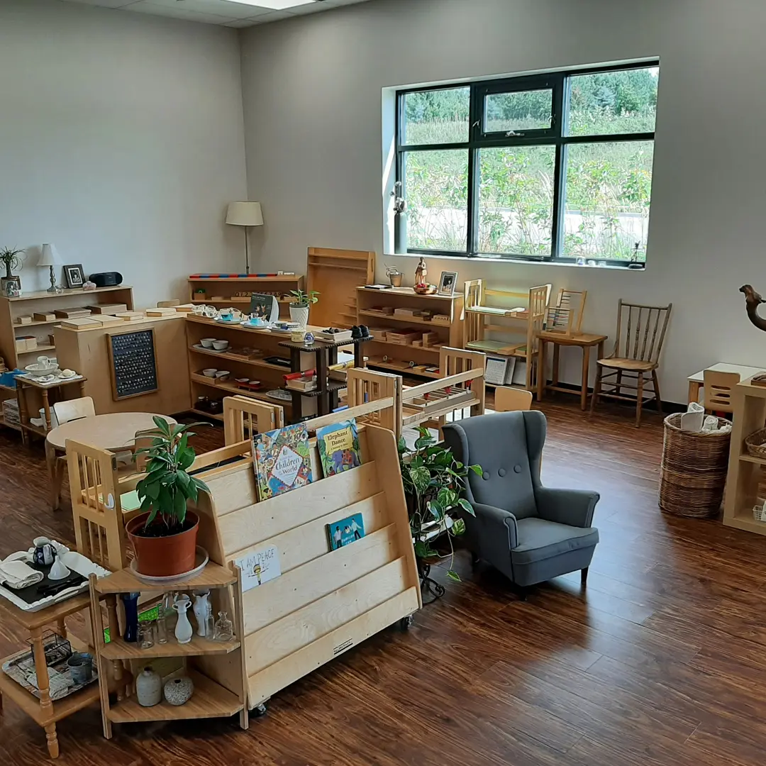 Bright, organized Montessori classroom with wooden furniture, educational materials, and a cozy chair; plants and books add a welcoming touch by a large window.