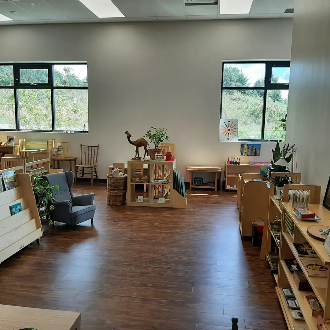 Bright classroom with wooden furniture, bookshelves, plants, and a camel sculpture. Large windows offer views of greenery outside. Cozy and organized learning space.