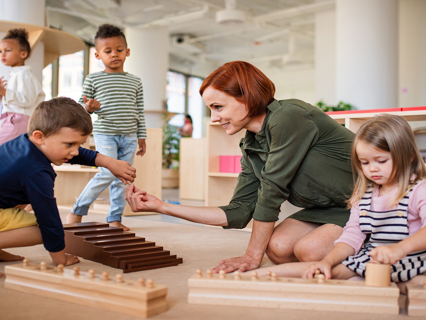 A person and children engage in educational activities using wooden blocks in a bright, modern classroom setting.