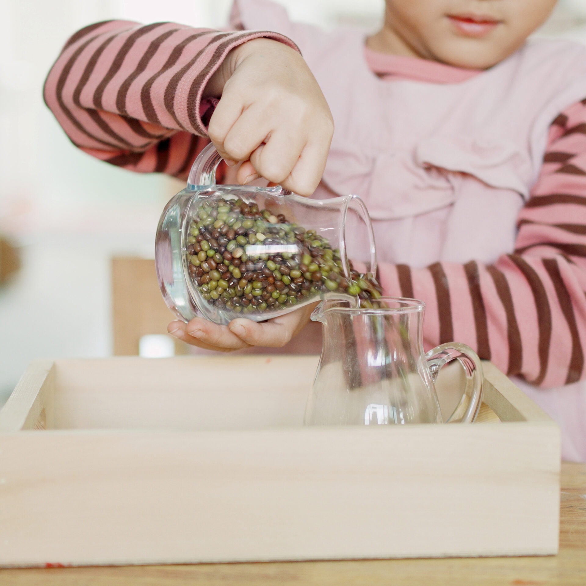 A child pours green beans from one glass pitcher to another within a wooden box, practicing coordination skills.
