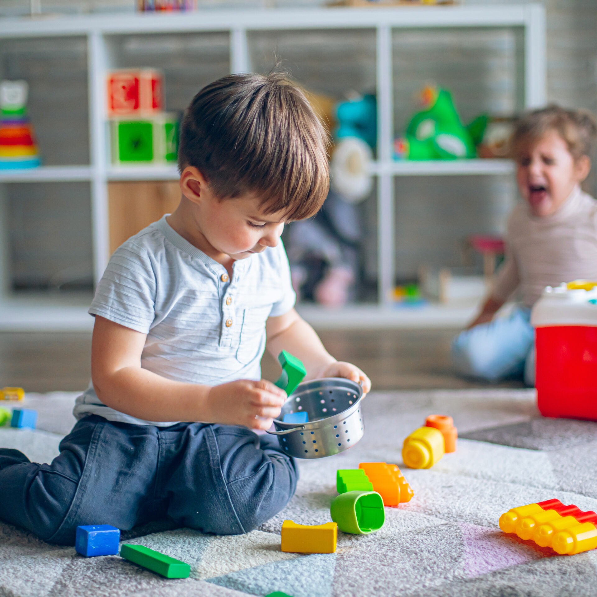 Two children are playing with colorful toys on a rug in a bright, organized room. One child focuses intently, other looks surprised.