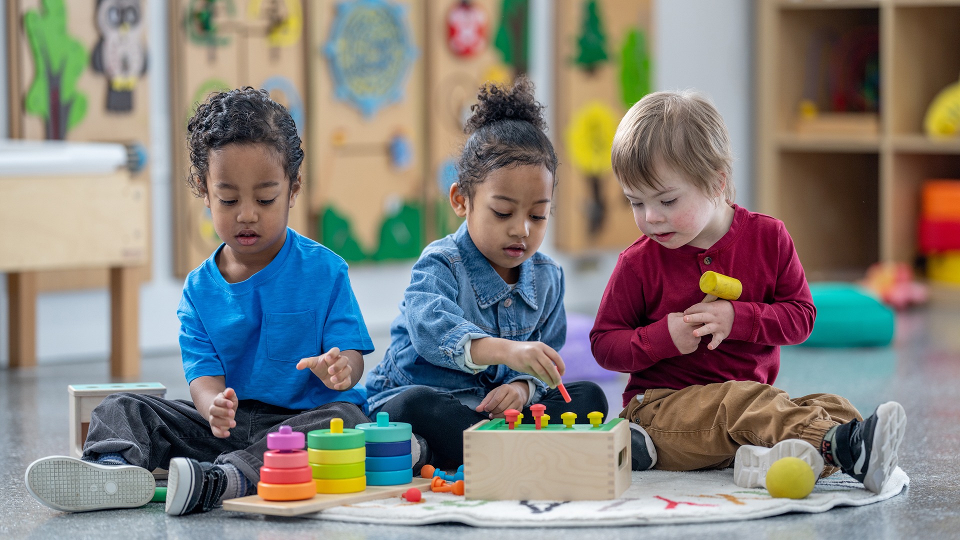 Three children are seated on the floor, playing with colorful educational toys in a bright and cheerful classroom setting.